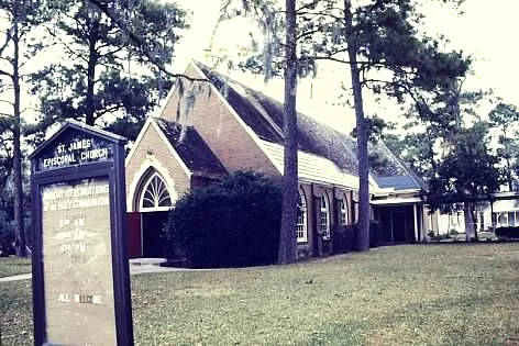 Church with a sign in front of it, surrounded by houses, trees, plants, and grass